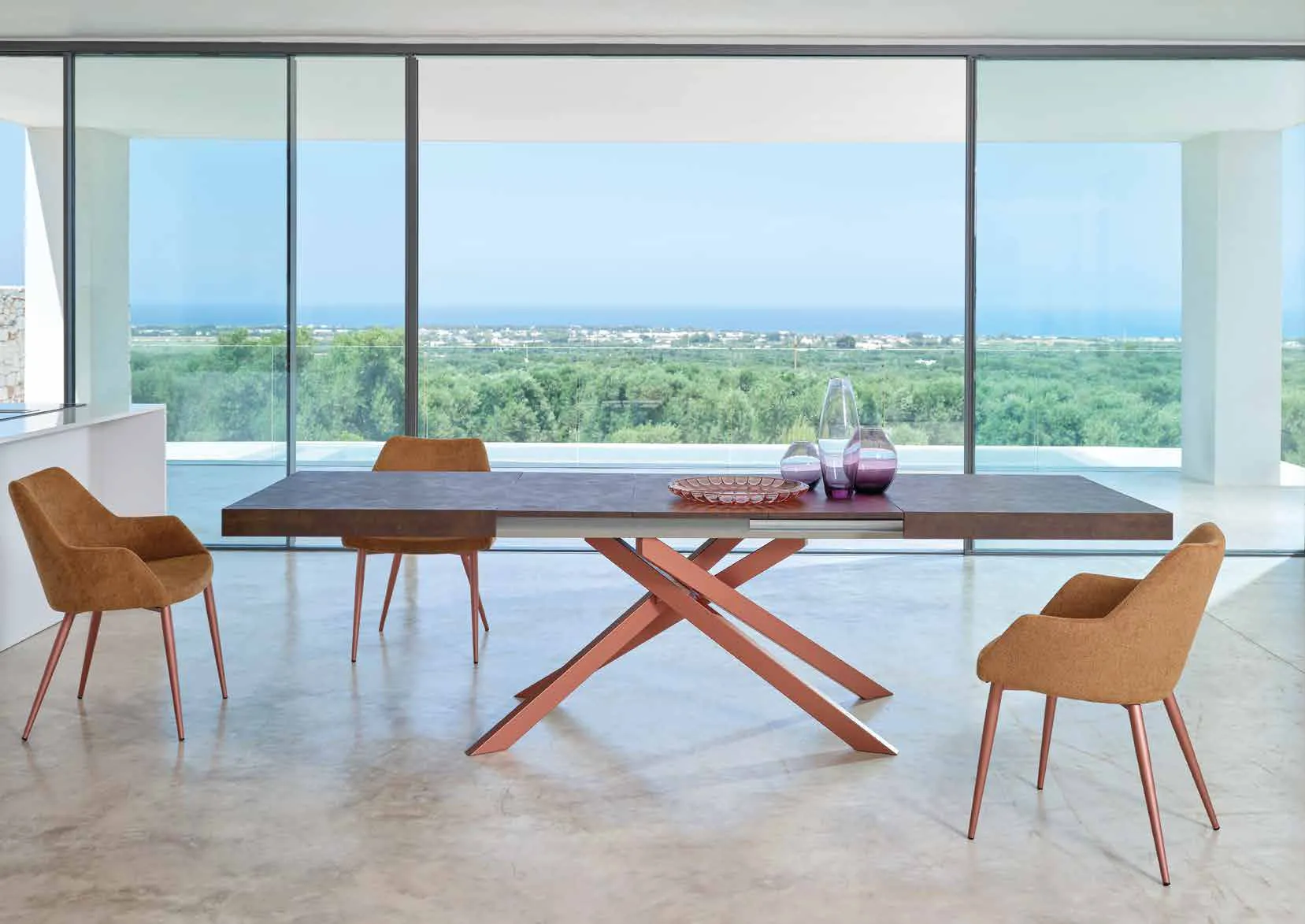 Tavolo Boston Friulsedie con gambe incrociate oro rosa e poltroncine arancio davanti a una vetrata sul mare