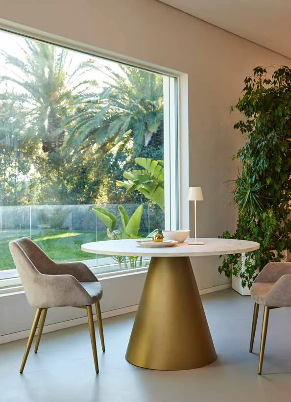 Tavolo Globe Friulsedie con base dorata e piano Tivoli Travertino davanti a una vetrata