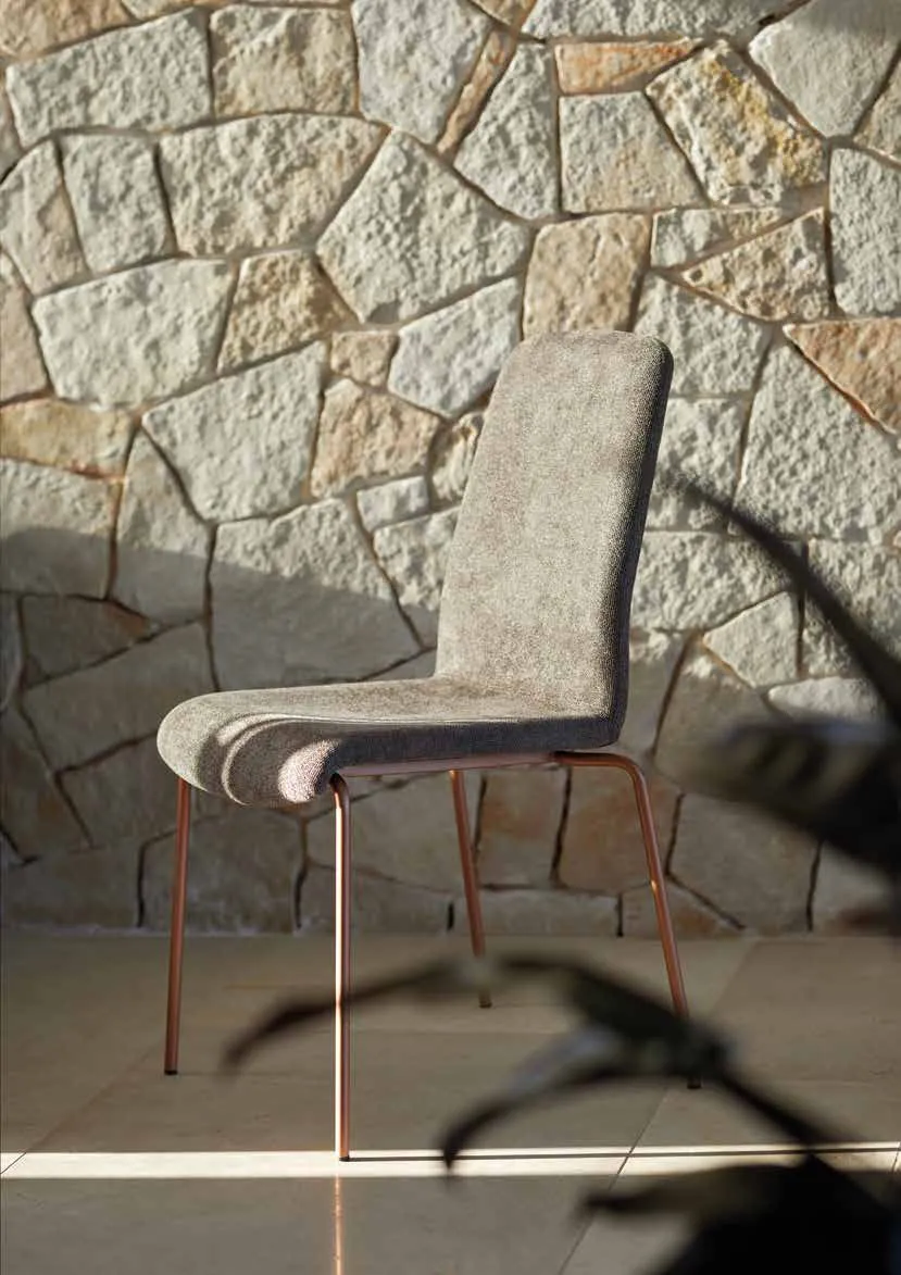 Sedia Mia Friulsedie in tessuto chiaro con gambe oro rosa davanti a un muro in pietra