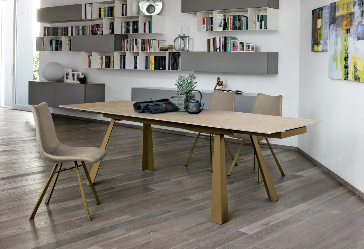 Tavolo Ponente Target Point con piano effetto legno e gambe dorate svasate in un soggiorno con libreria
