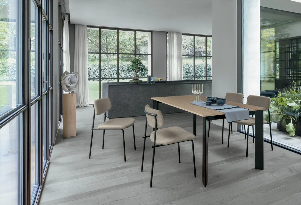 Tavolo Saturno Target Point con piano effetto legno e gambe nere, sedie beige accanto alla vetrata