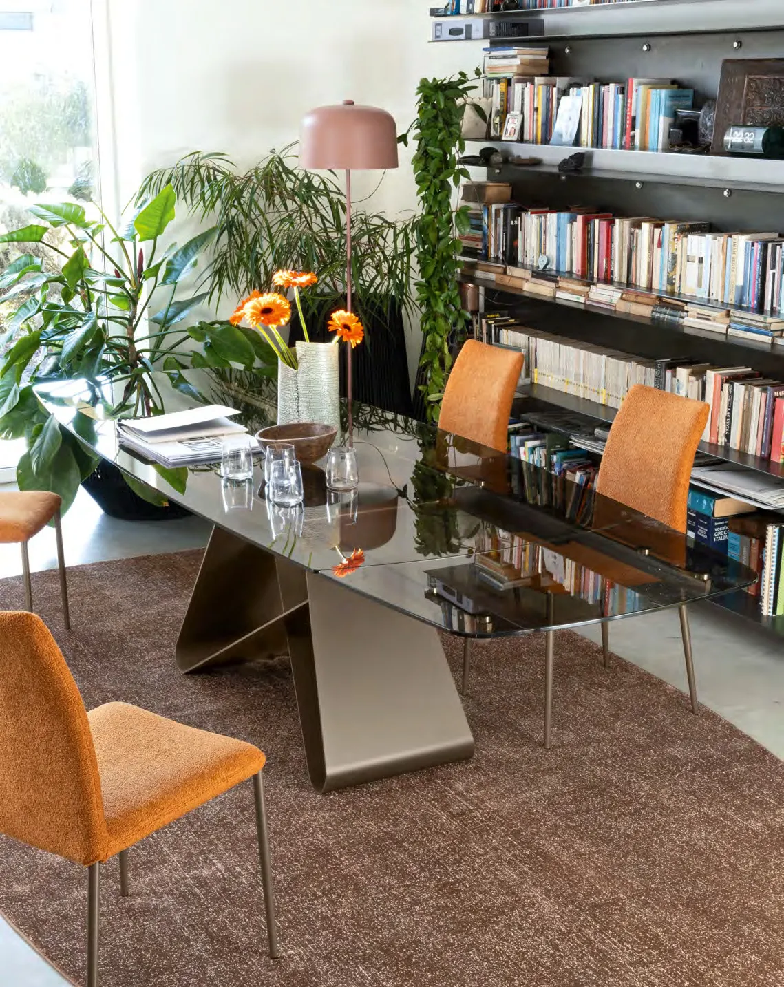 Tavolo Gemini Connubia sagomato con piano in vetro bronzo e base lava davanti a una libreria, con sedie arancio