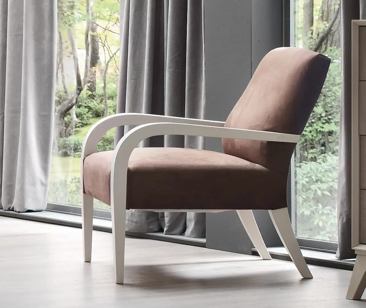 Poltroncina Suite Le Fablier con braccioli bianchi e rivestimento marrone davanti a una vetrata