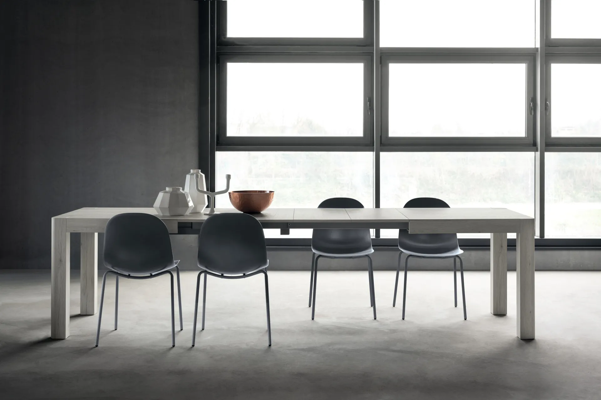 Tavolo Winter Scavolini bianco allungato con sedie scure davanti a una vetrata industriale
