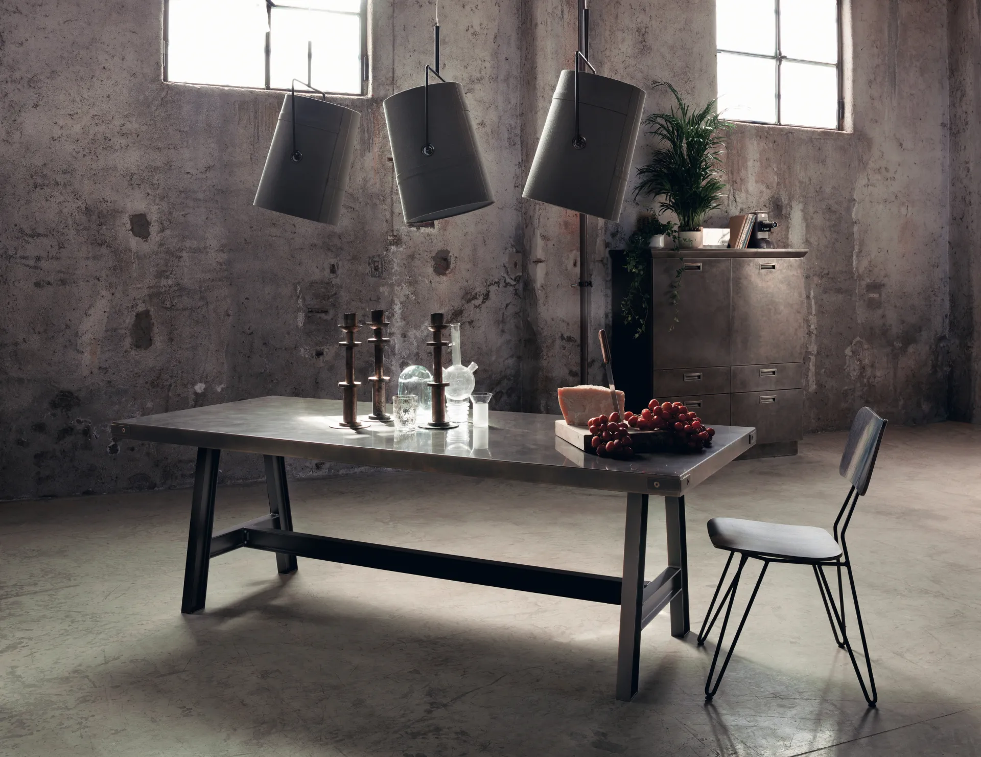 Misfit Table Scavolini con cavalletti in metallo scuro, piano grigio e sedia in filo in un ambiente industriale