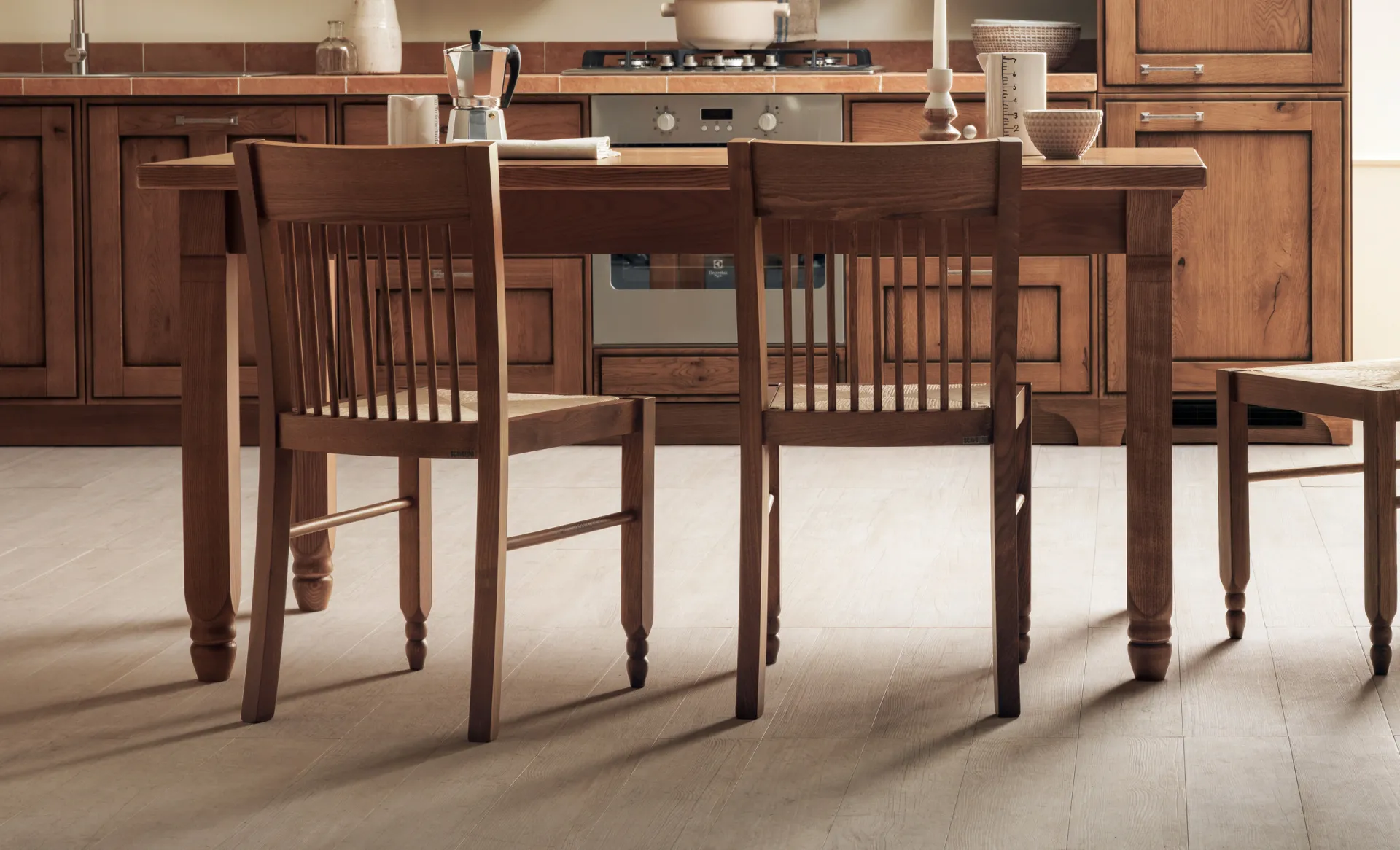 Tavolo Gea Scavolini in legno con gambe tornite e sedie impagliate davanti a una cucina rustica