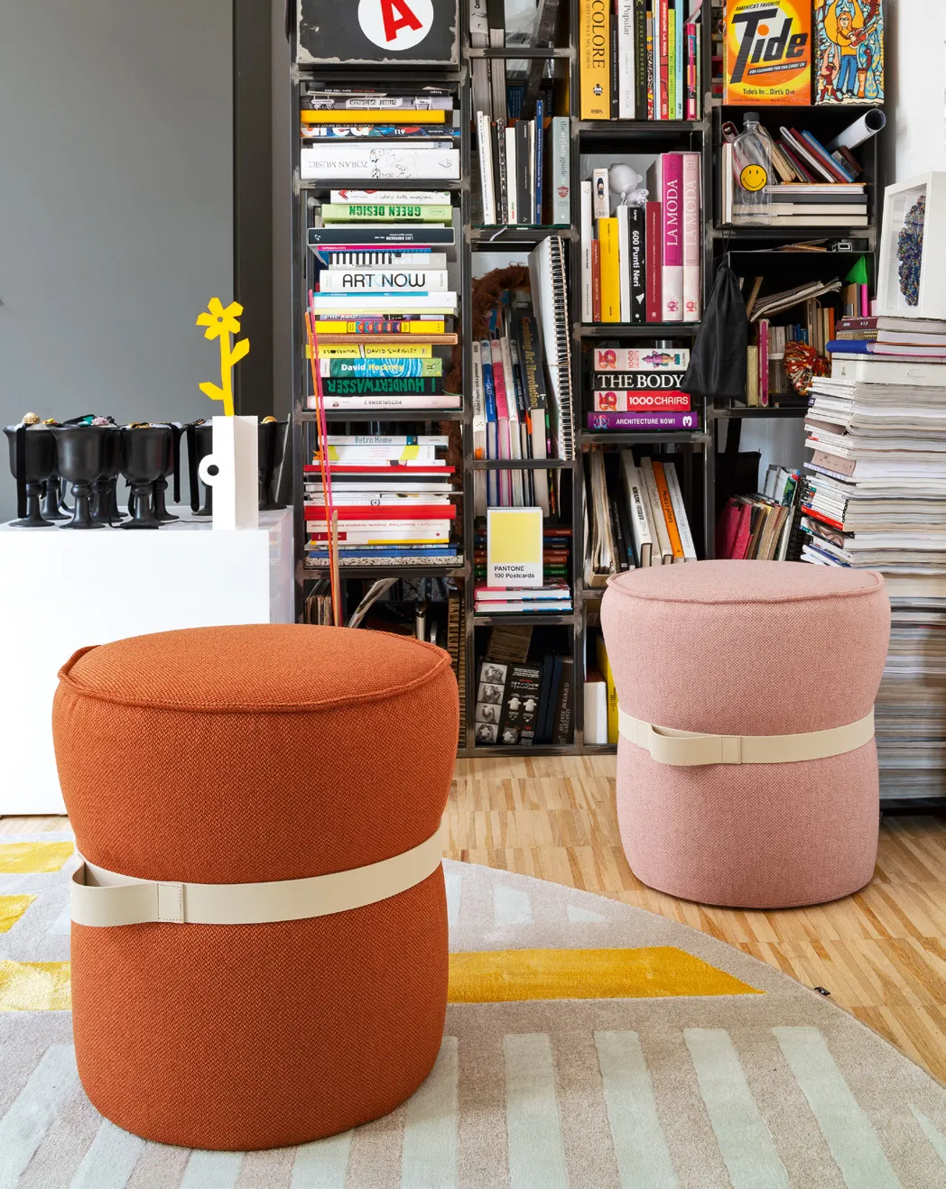Pouf Pof Connubia in tessuto zafferano e rosa con cinghia chiara davanti a una libreria colma