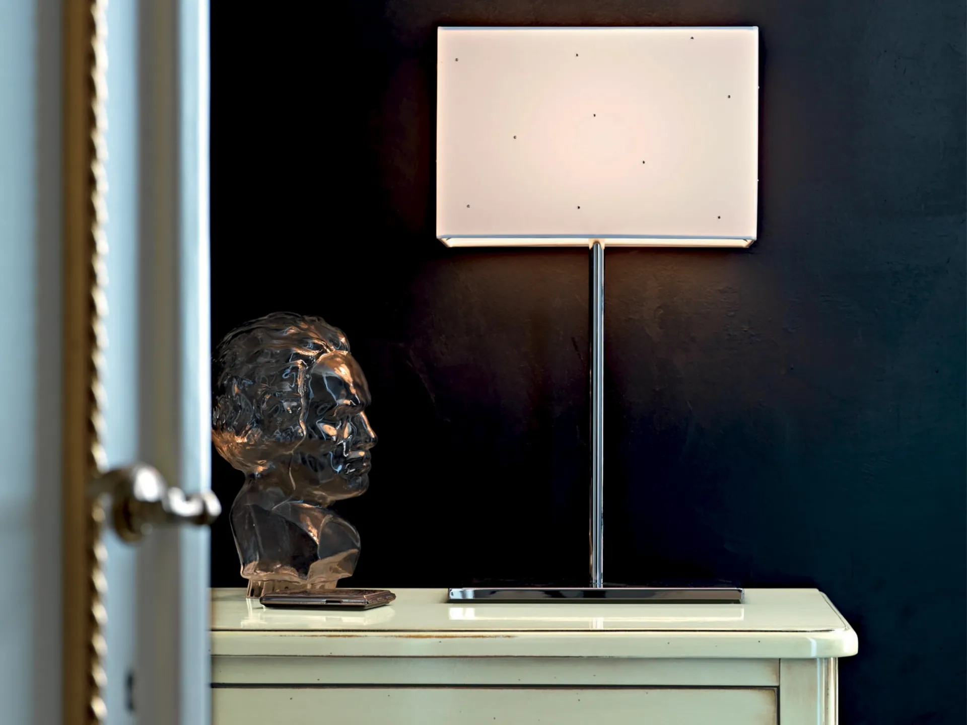 Lampada Glam Le Fablier accesa su un mobile bianco accanto a una scultura