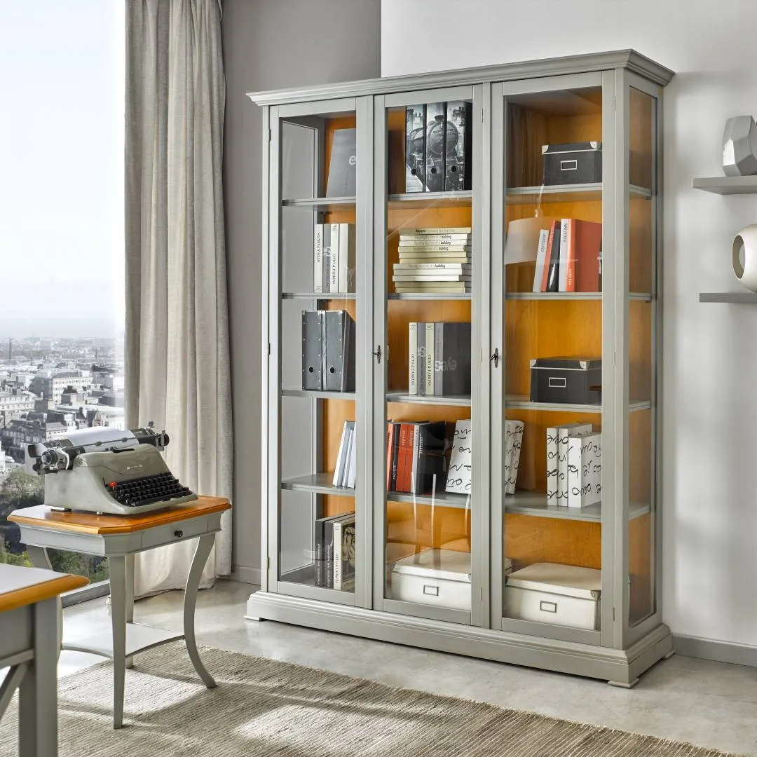 Libreria a tre ante vetrate Tosato 60.25 grigia con interni color miele, accanto a uno scrittoio