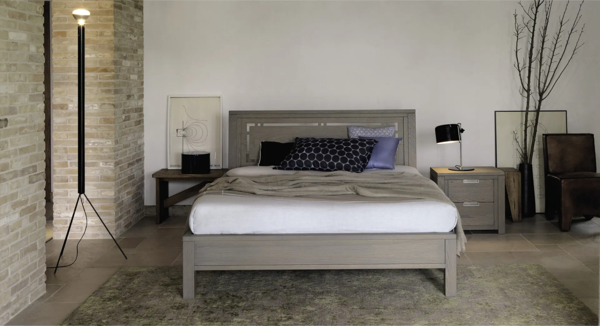 Letto Quadra Devina Nais color cenere con testiera riquadrata in una camera con parete in pietra
