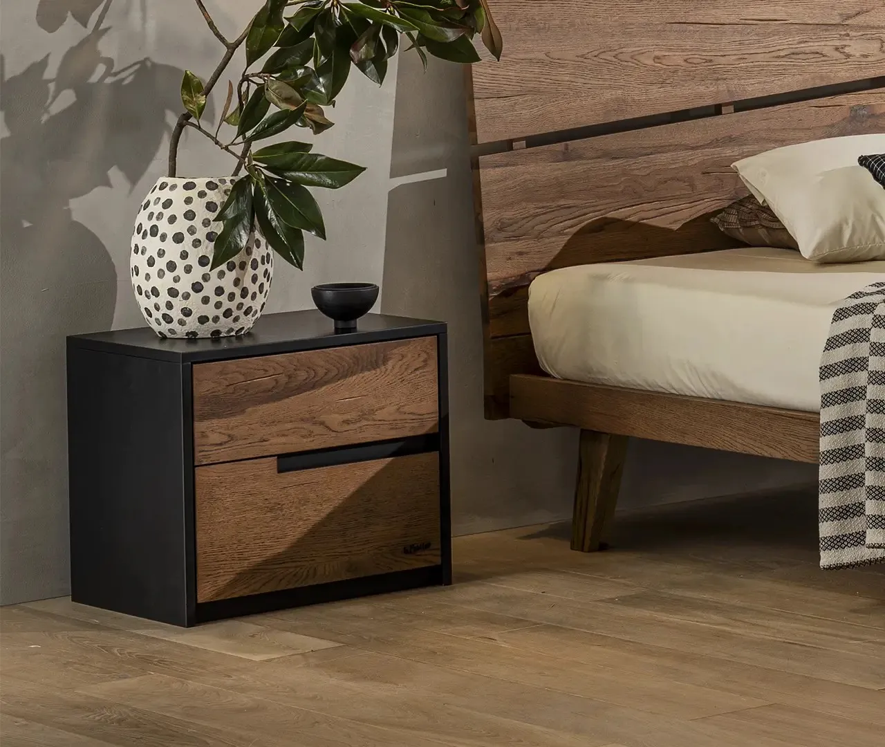 Comodino Salisburgo Le Fablier con due cassetti in rovere e struttura nera accanto a un letto in legno