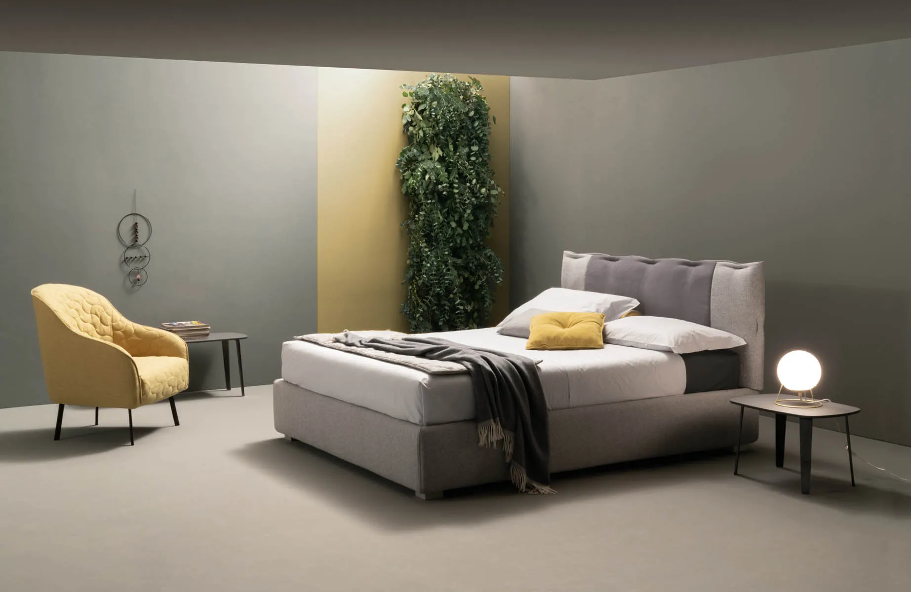 Letto imbottito Dress Samoa grigio con pannello scuro sulla testiera, accanto a una poltroncina gialla
