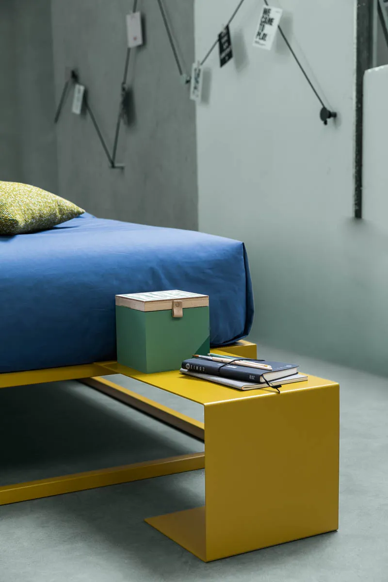 Comodino Frame Samoa giallo accanto a un letto con copriletto blu