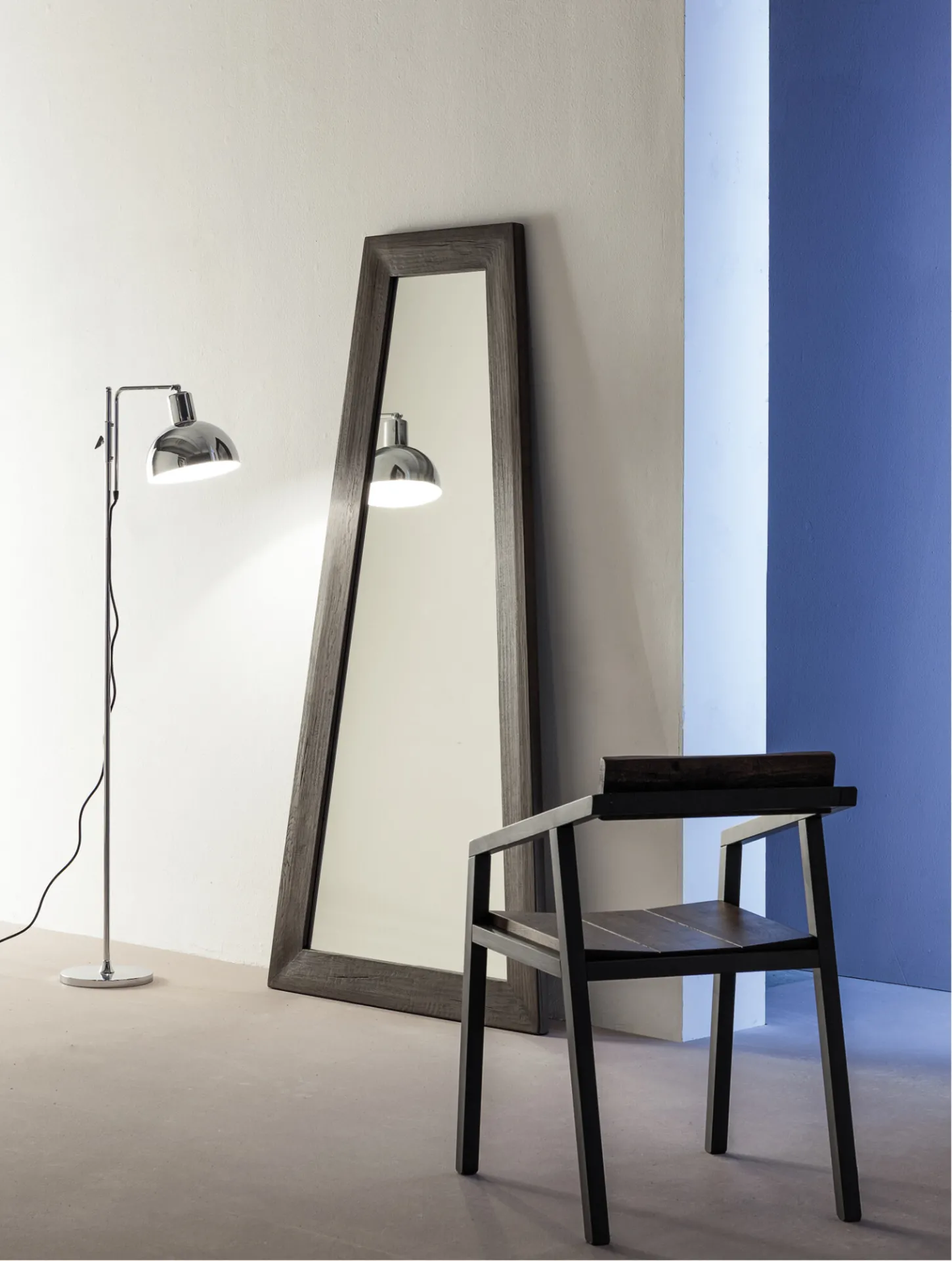 Specchio Trapezio Devina Nais con cornice scura appoggiato alla parete vicino a una sedia