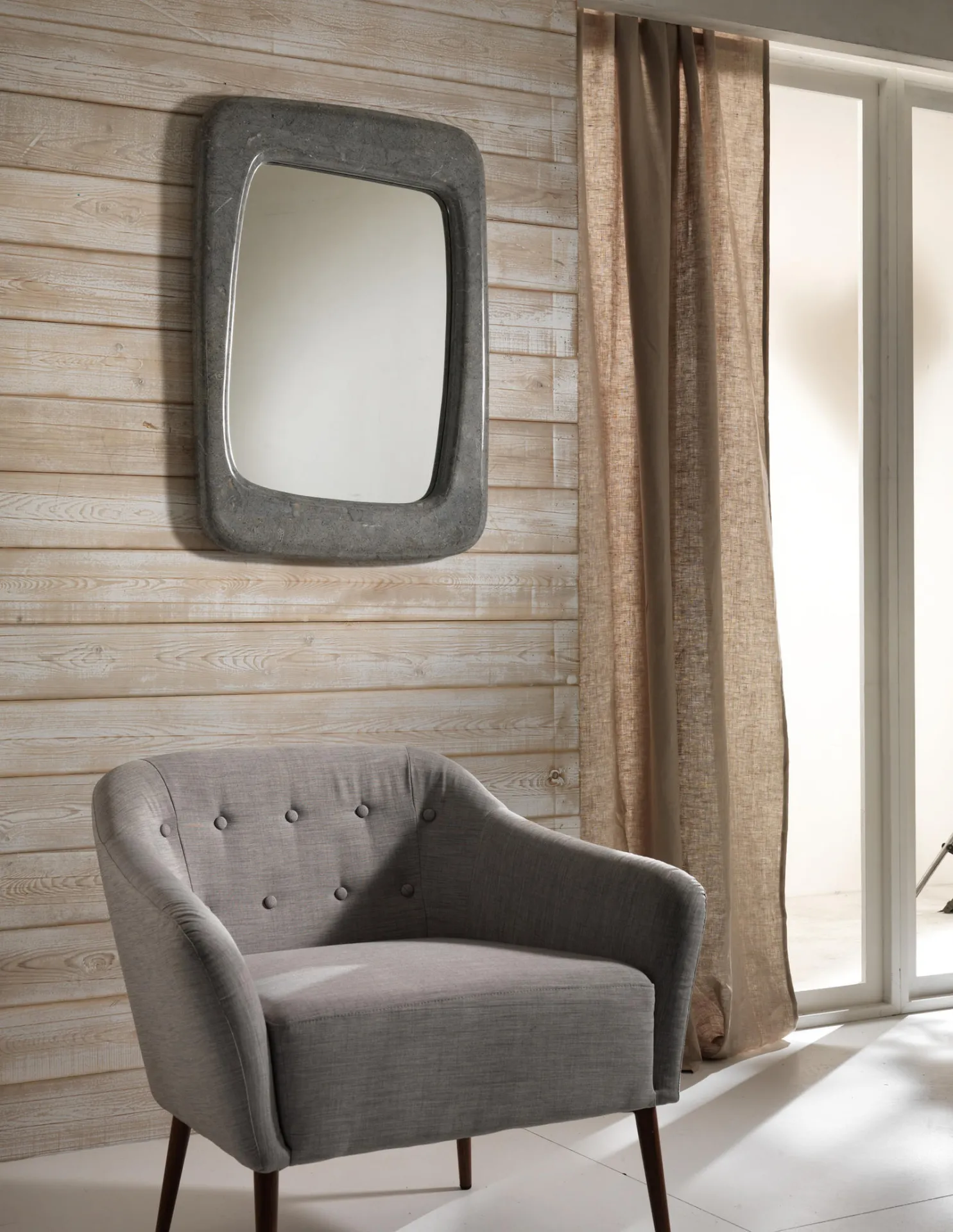 Specchio Smooth Stones grigio su una parete in legno accanto a una poltrona