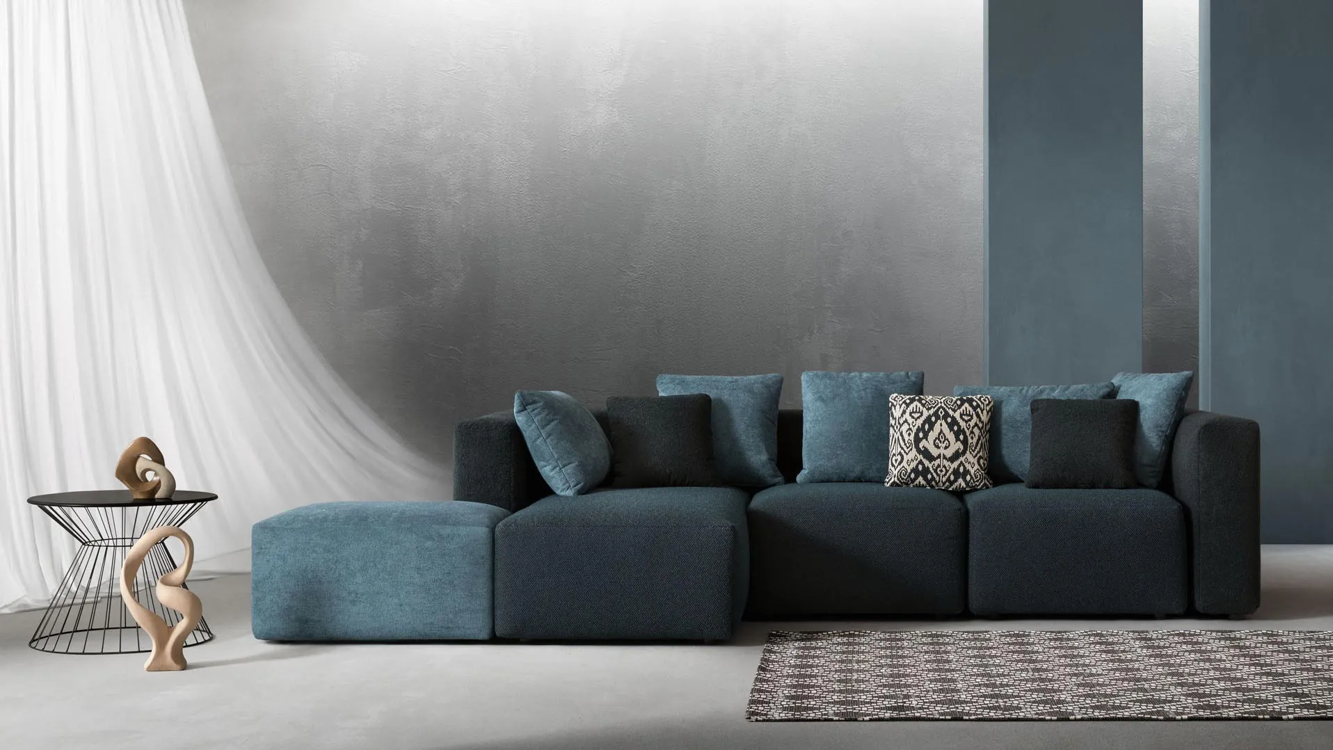 Divano modulare Sassy Samoa blu petrolio con pouf, a blocchi squadrati, davanti a una parete grigia