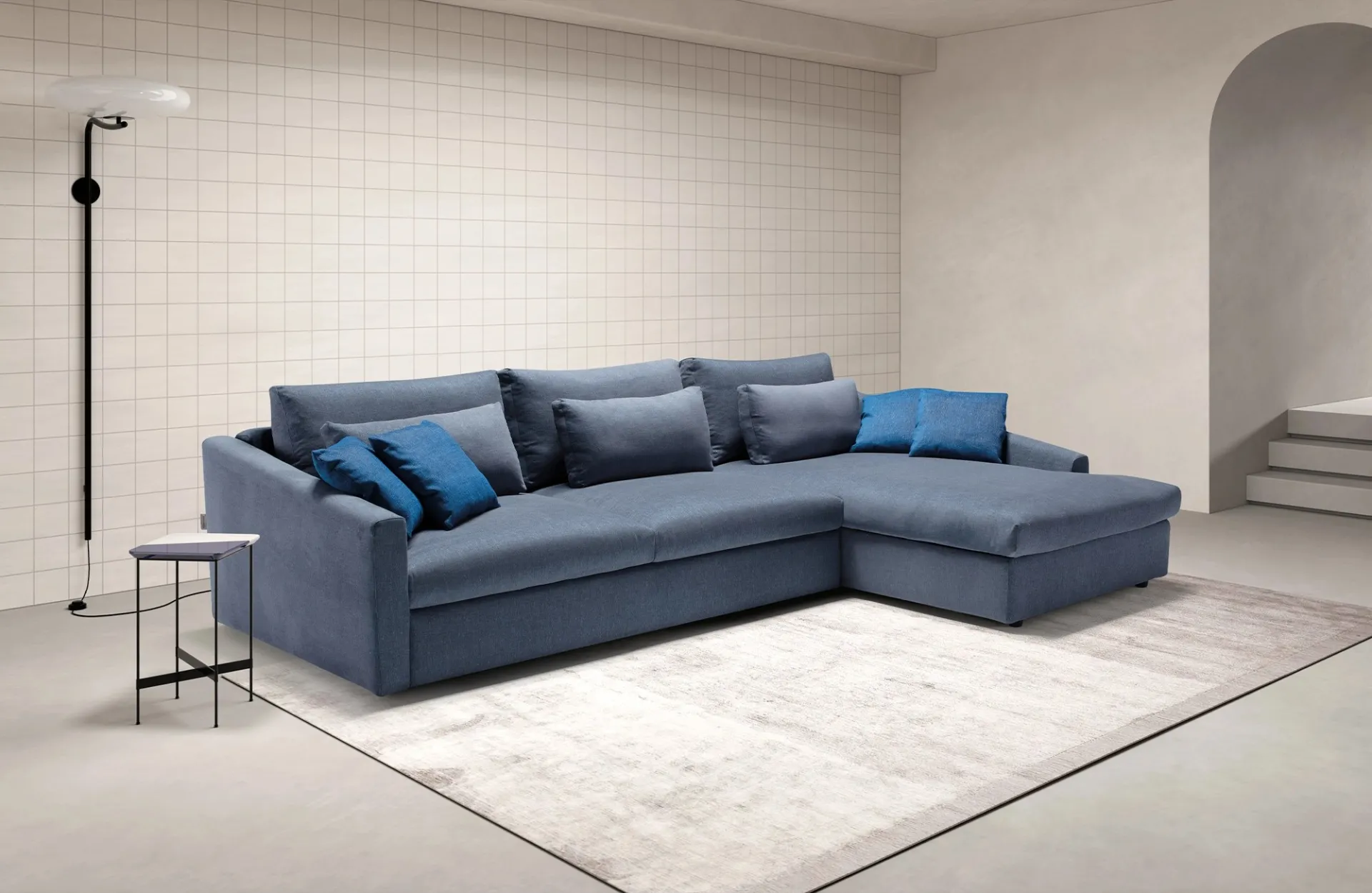 Divano letto Mithos Dienne azzurro con chaise longue e cuscini blu in una stanza piastrellata