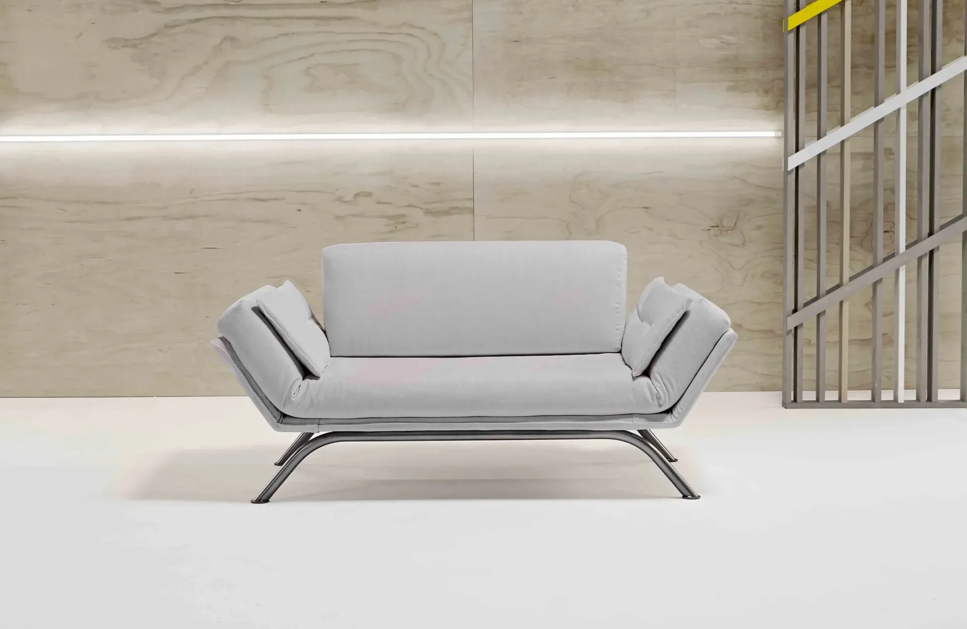 Divano letto Nicla Dienne grigio chiaro con braccioli ad ala su base metallica davanti a una parete in legno