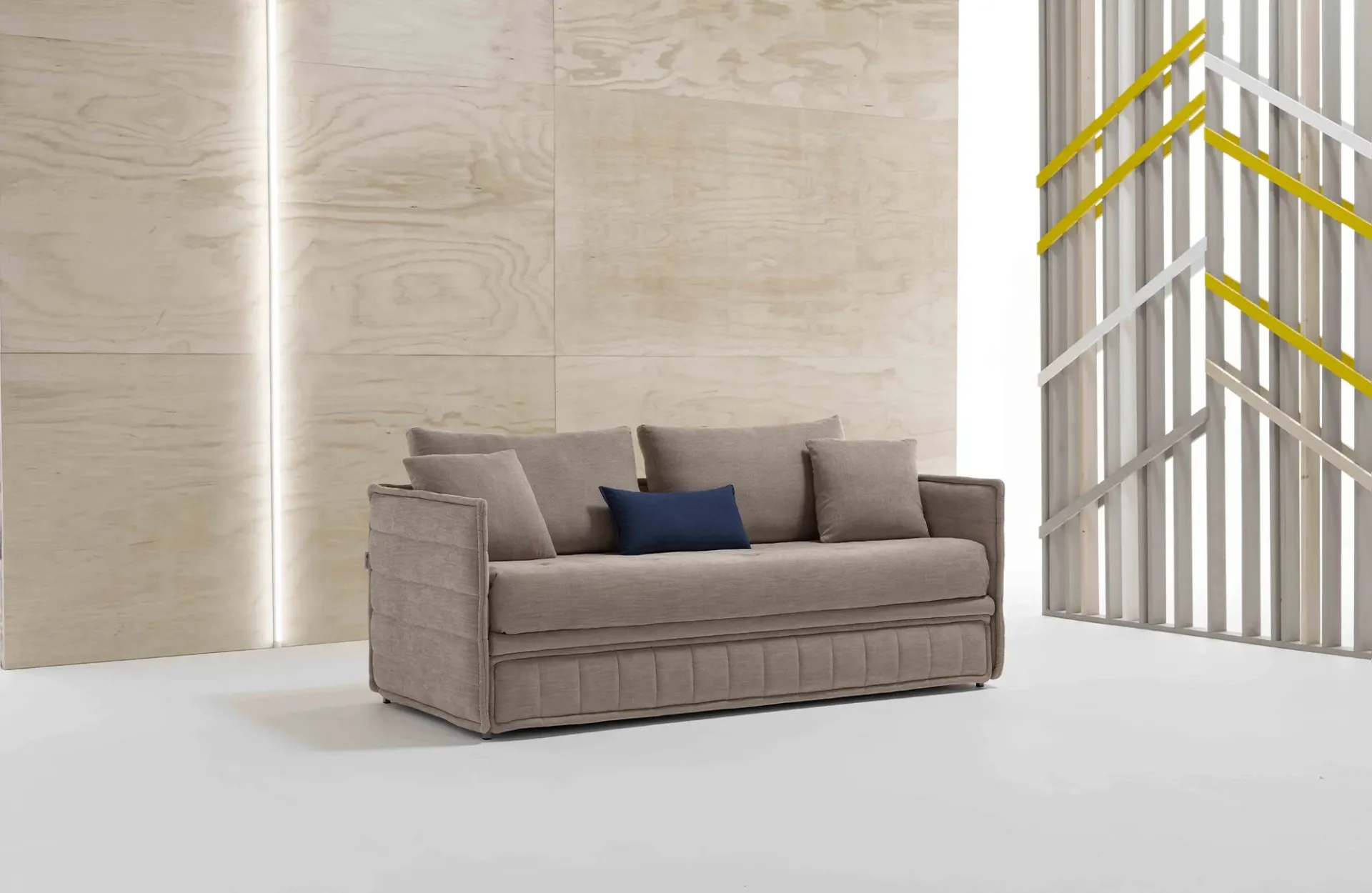 Divano letto Jolie Dienne color sabbia con fianchi trapuntati e cuscino blu davanti a una parete in legno