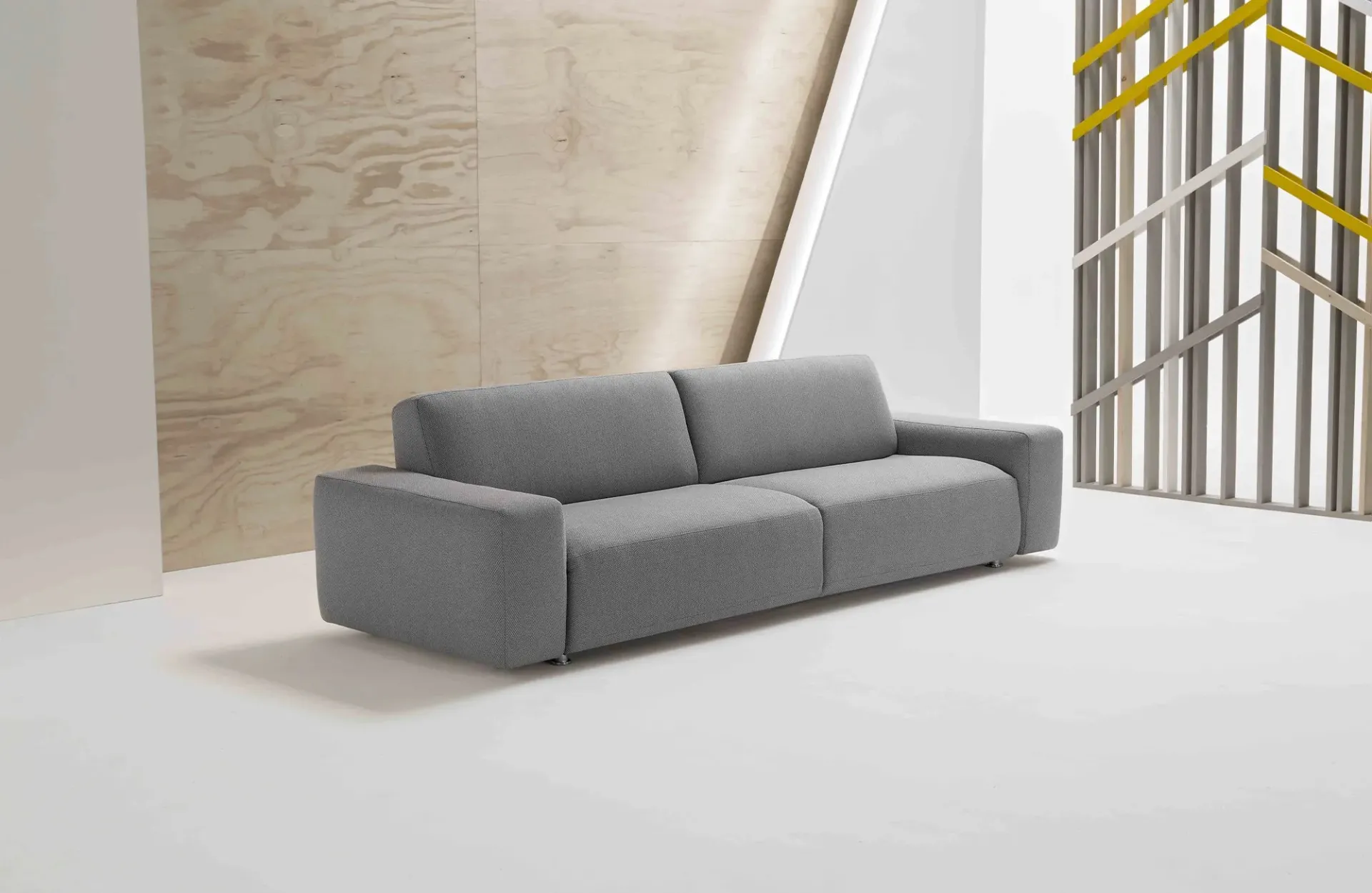 Divano letto Cross Dienne grigio con braccioli a blocco davanti a una parete in legno chiaro