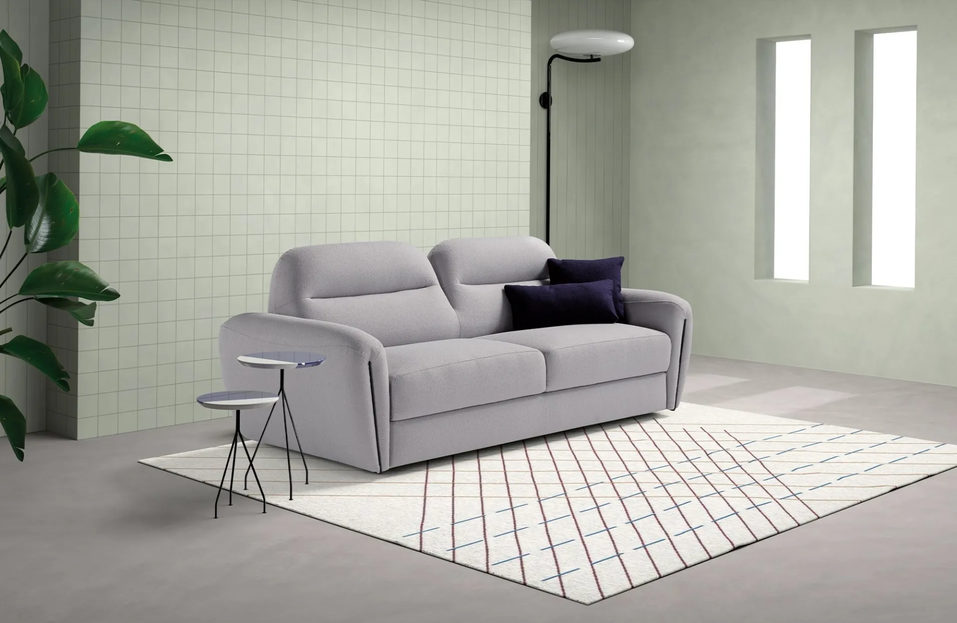 Divano letto Soul Dienne grigio perla con cuscini blu scuro in una stanza con piastrelle e pianta
