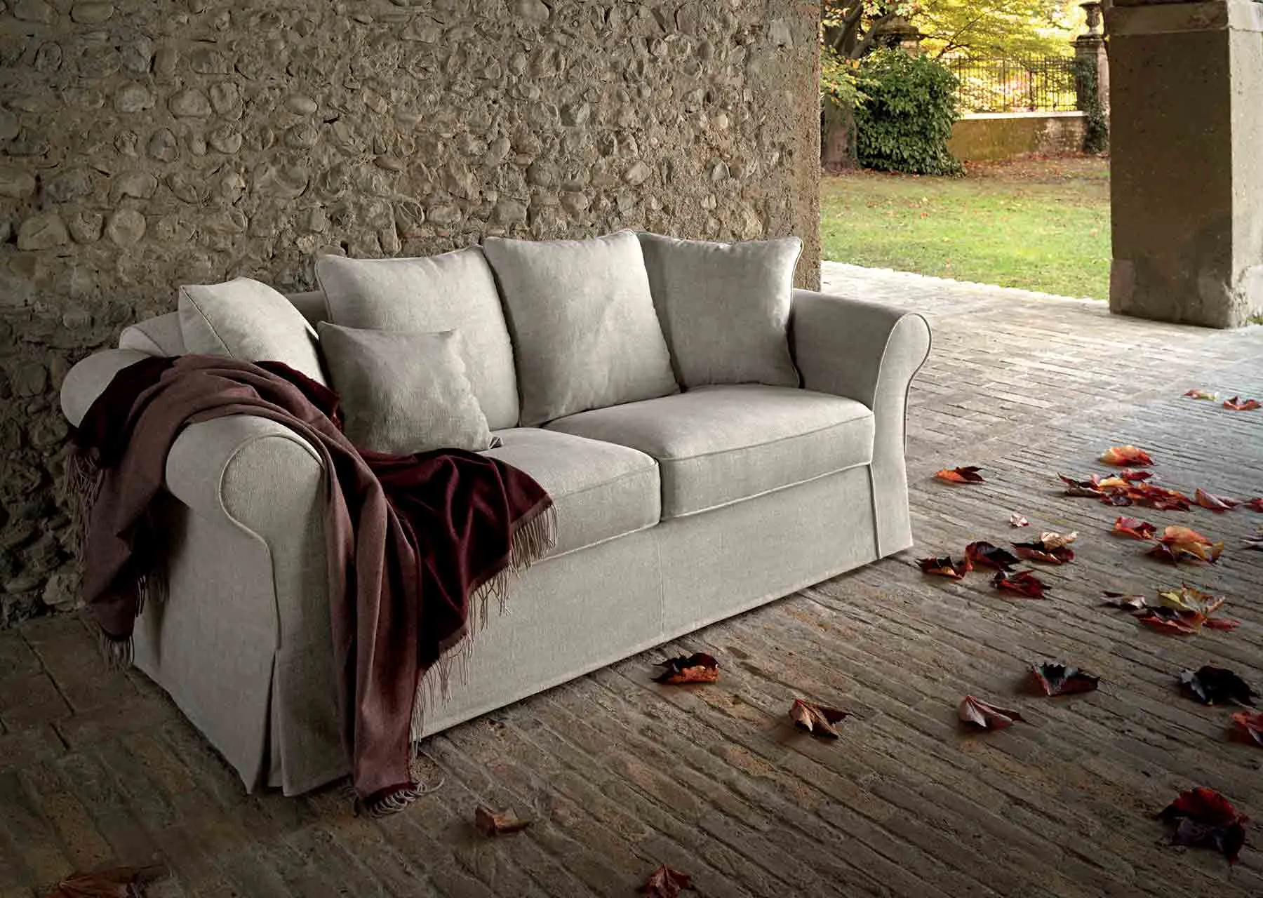 Divano classico Elite Samoa tre posti in tessuto chiaro con plaid bordeaux, in un cortile lastricato