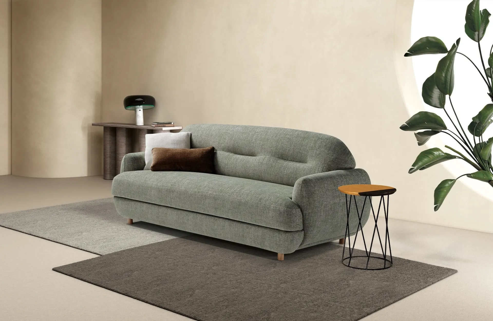 Divano letto Plum Dienne verde salvia con cuscini marroni, tra un tavolino in metallo e una pianta