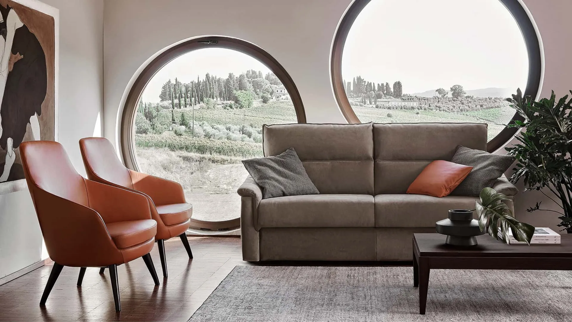 Divano letto Bormio Rosini grigio con poltrone cuoio davanti a finestre tonde