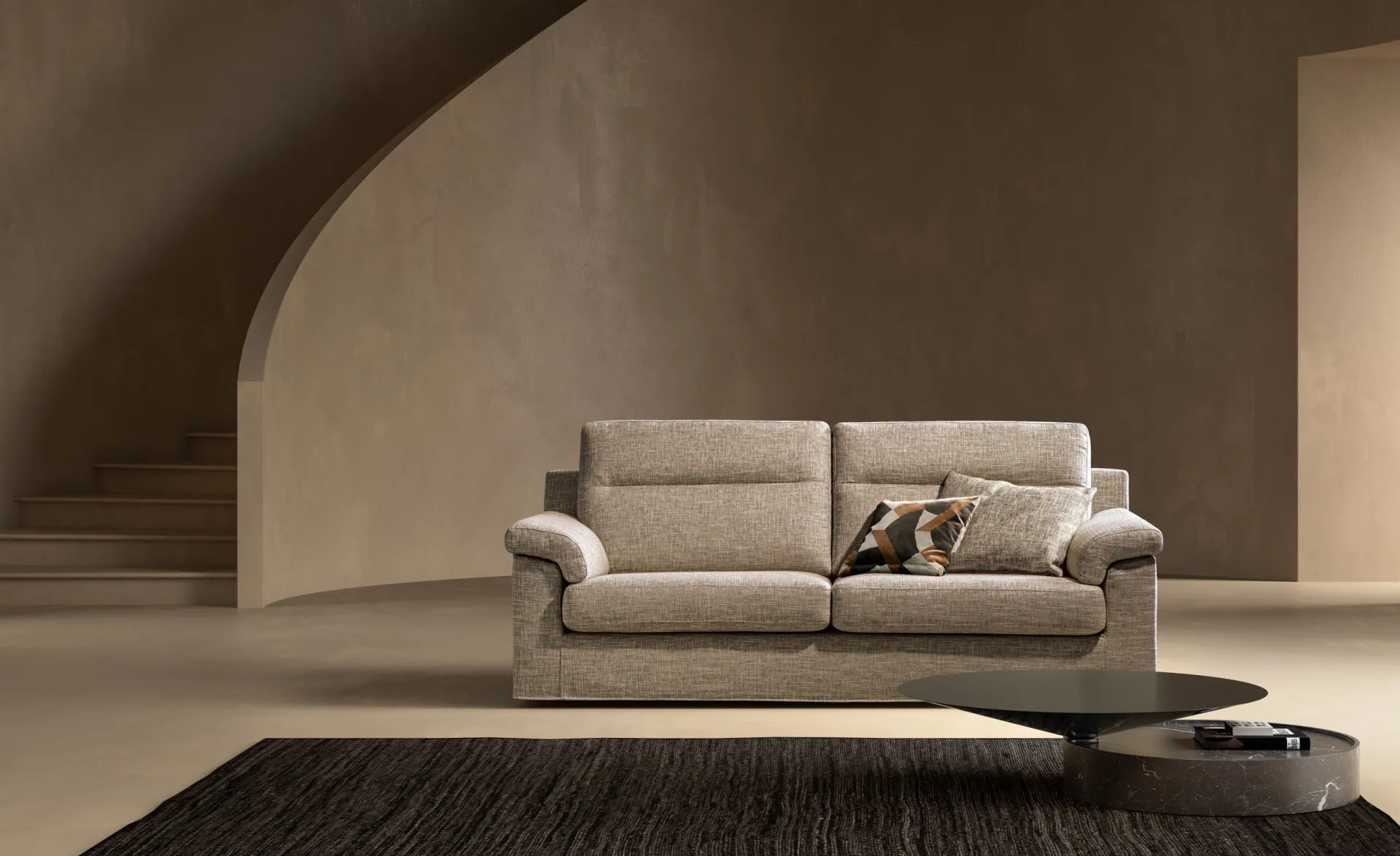Divano Skill Samoa beige a tre posti con cuscini a fantasia, sotto una scala curva