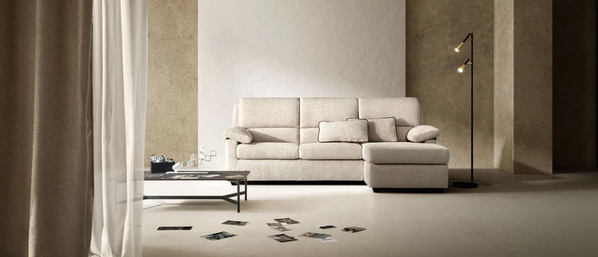 Divano Sun Samoa chiaro con chaise longue, fra tende e una lampada da terra