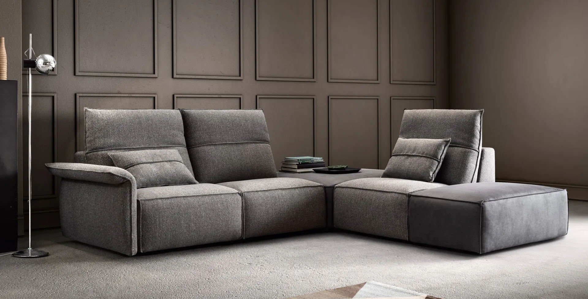 Divano Deep Vibe Samoa grigio ad angolo con poggiatesta alzati, accanto a una lampada