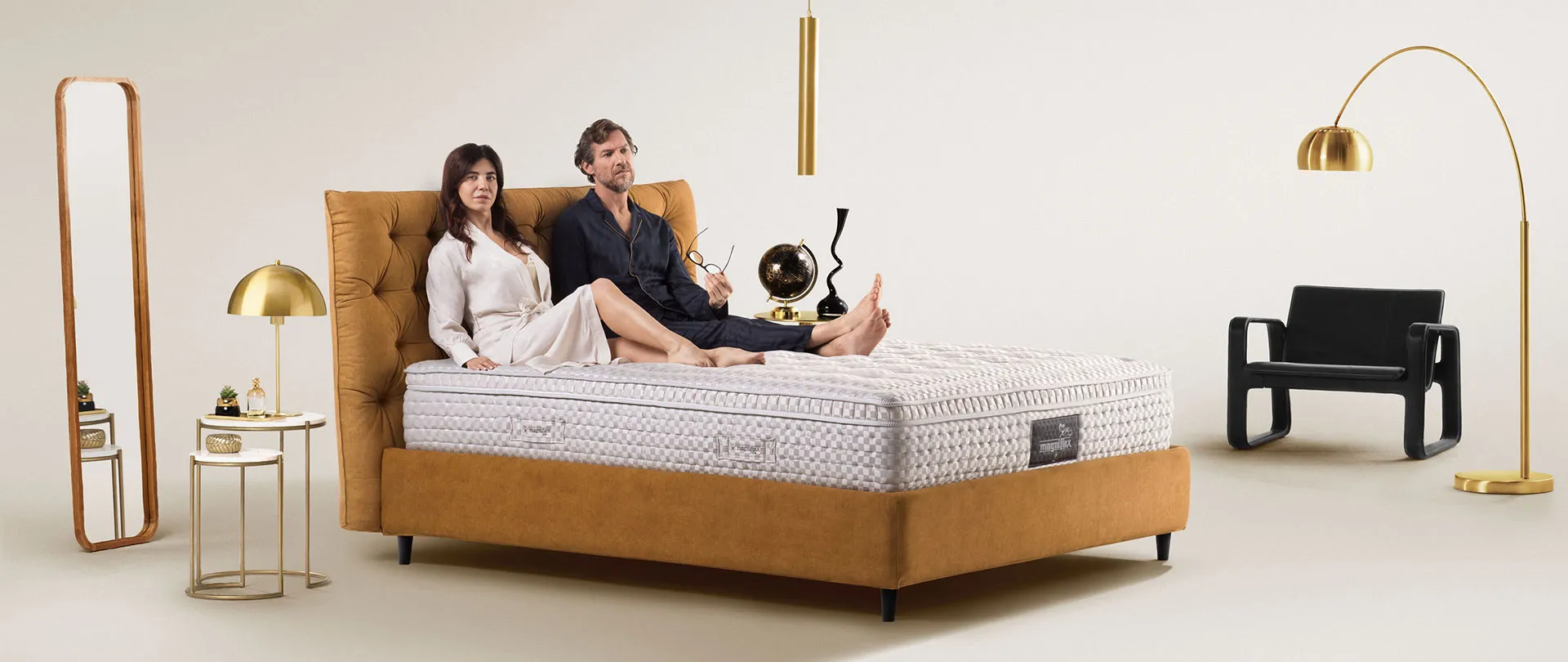 Materasso della collezione Magnifico Magniflex su letto capitonné color ocra, con una coppia seduta contro la testiera
